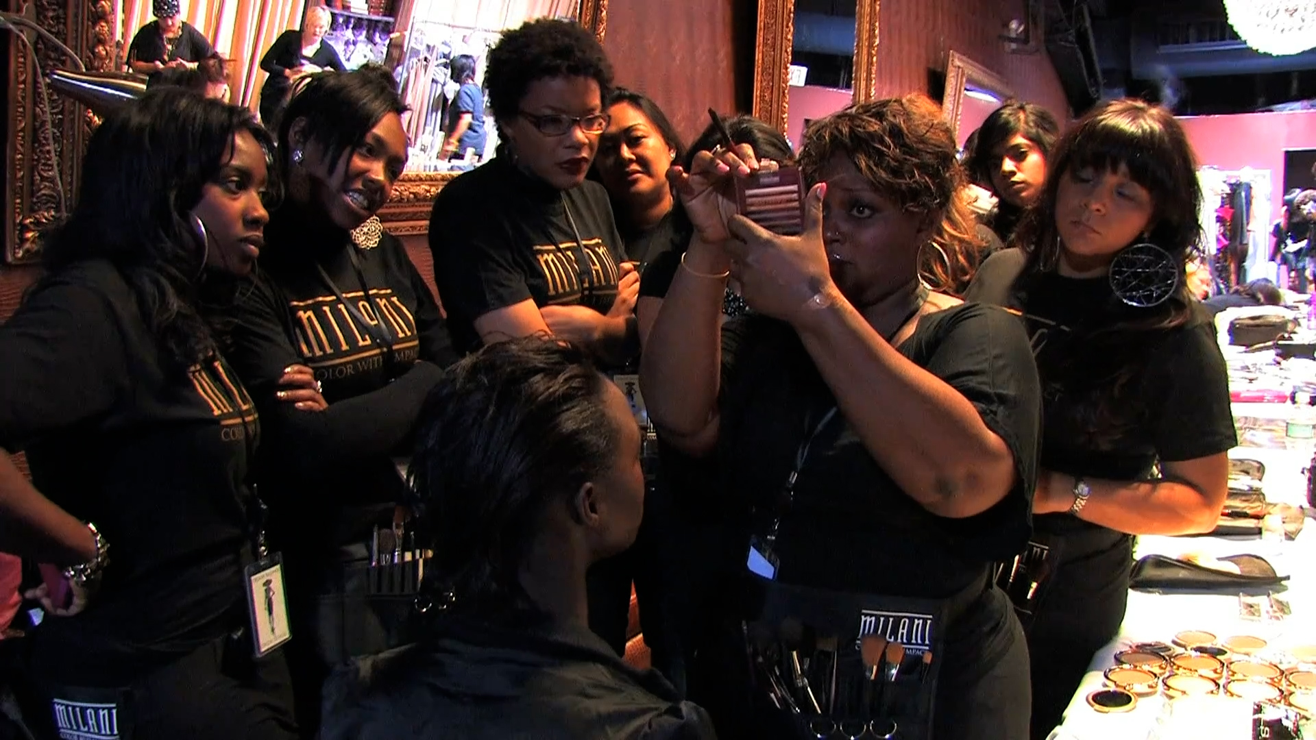Ashunta Sheriff working her magic,  doing hair, makeup and manicures inside an upscale beauty salon. The scene is being filmed on video with a full shot from behind them as they stand next to tables filled with cosmetics, makeup brushes, scissors and nail polish scattered everywhere.