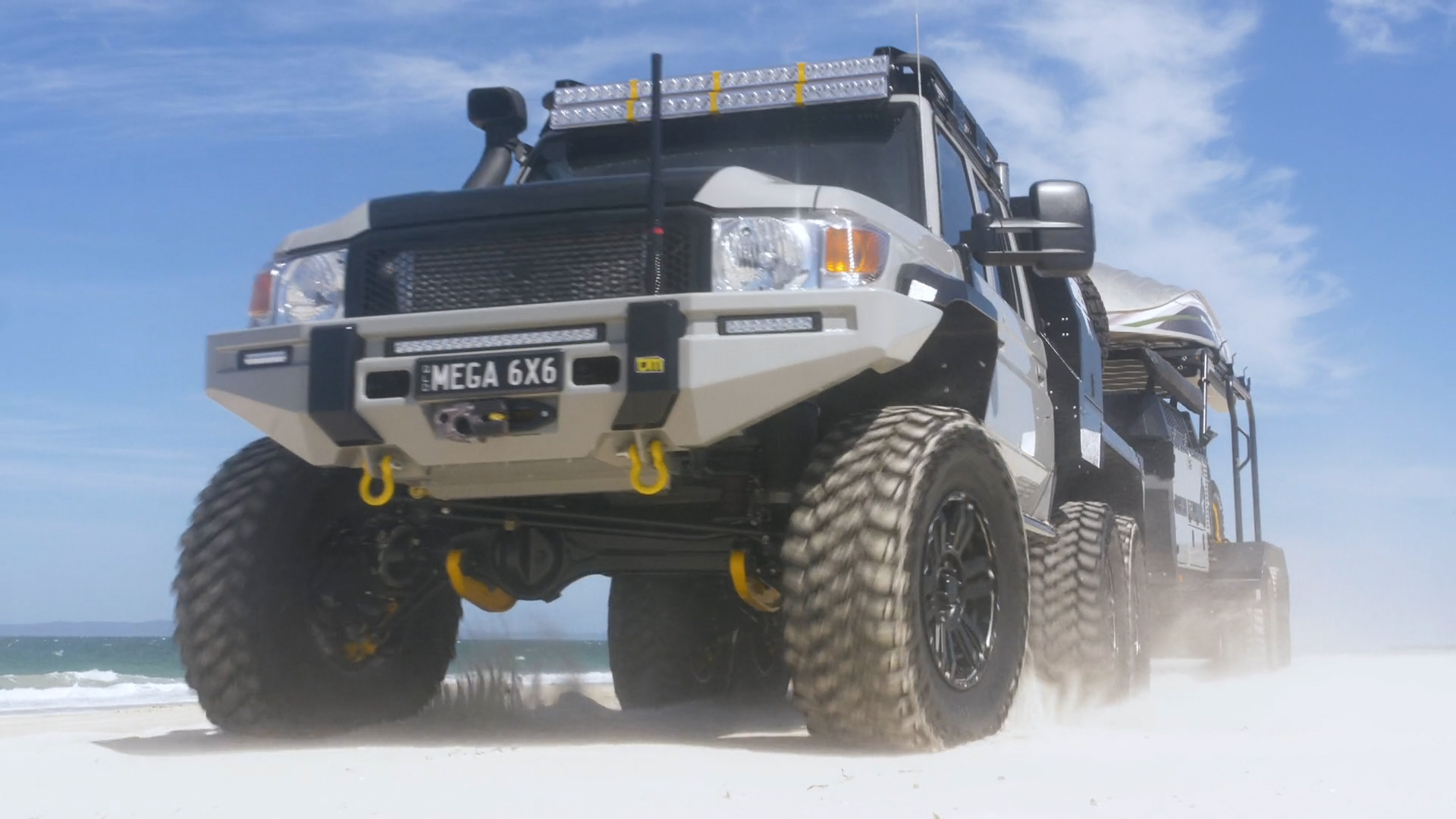 A wide angle photo of a super lifting off road vehicle with large wheels and very high headlights, on the beach in Australia. The truck is white and grey with yellow accents. It has black steel bodywork and no windows. There's dust flying around as it lift car, it also carries two rows of lights over its hood. In front there’s one rectangular light bar illuminating under every single wheel. A glass roof box sits above all the features.